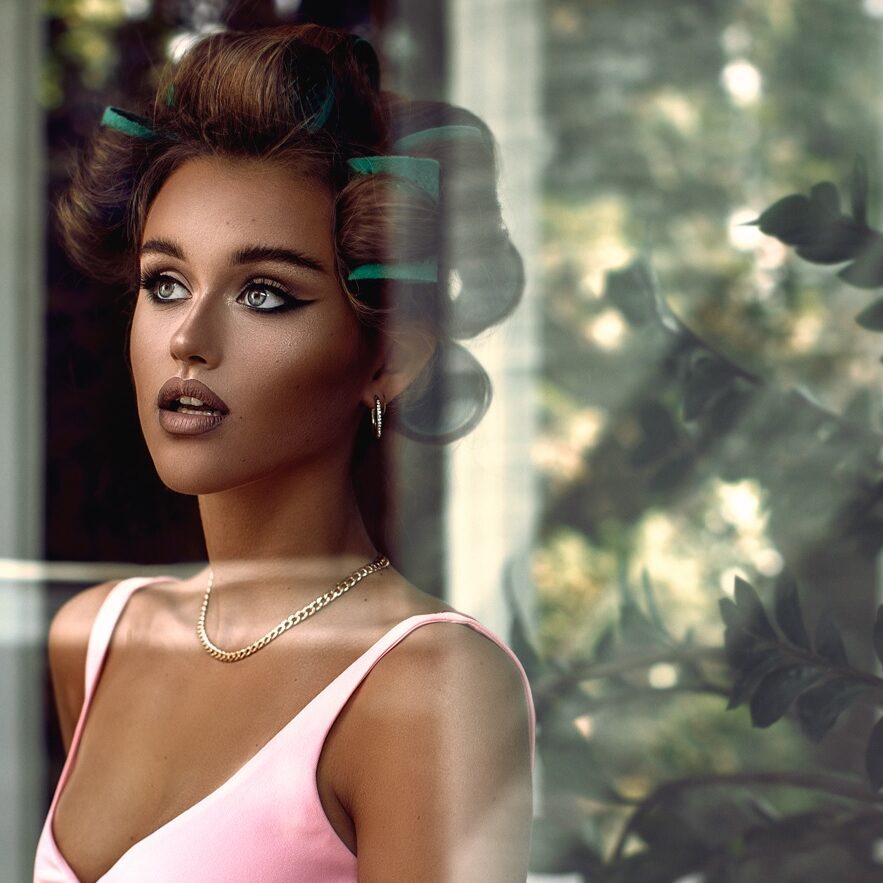 Model med eyeliner og ruller i håret foran et vindue i soft, cinematic lys