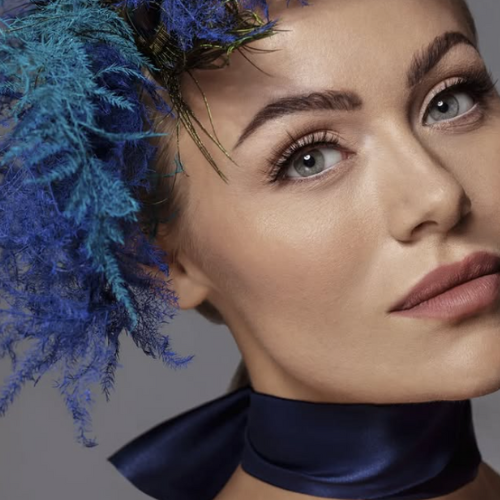 Elegant close-up af kvinde med soft glam makeup, markerede bryn og blå fjer headpiece.