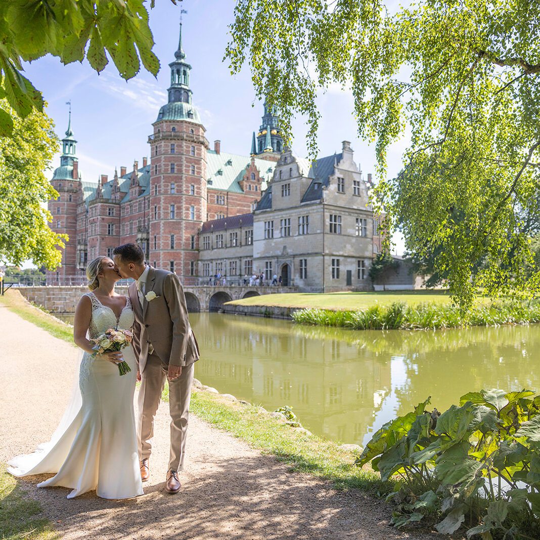 Brudepar der kysser i sollys ved sø og historisk slot med elegant brudemakeup og klassisk brudehår.