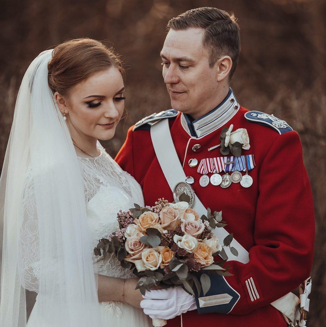 Brudepar udendørs; bruden med elegant opsat hår, naturlig brudemakeup og langt slør står tæt ved partner i rød uniform.