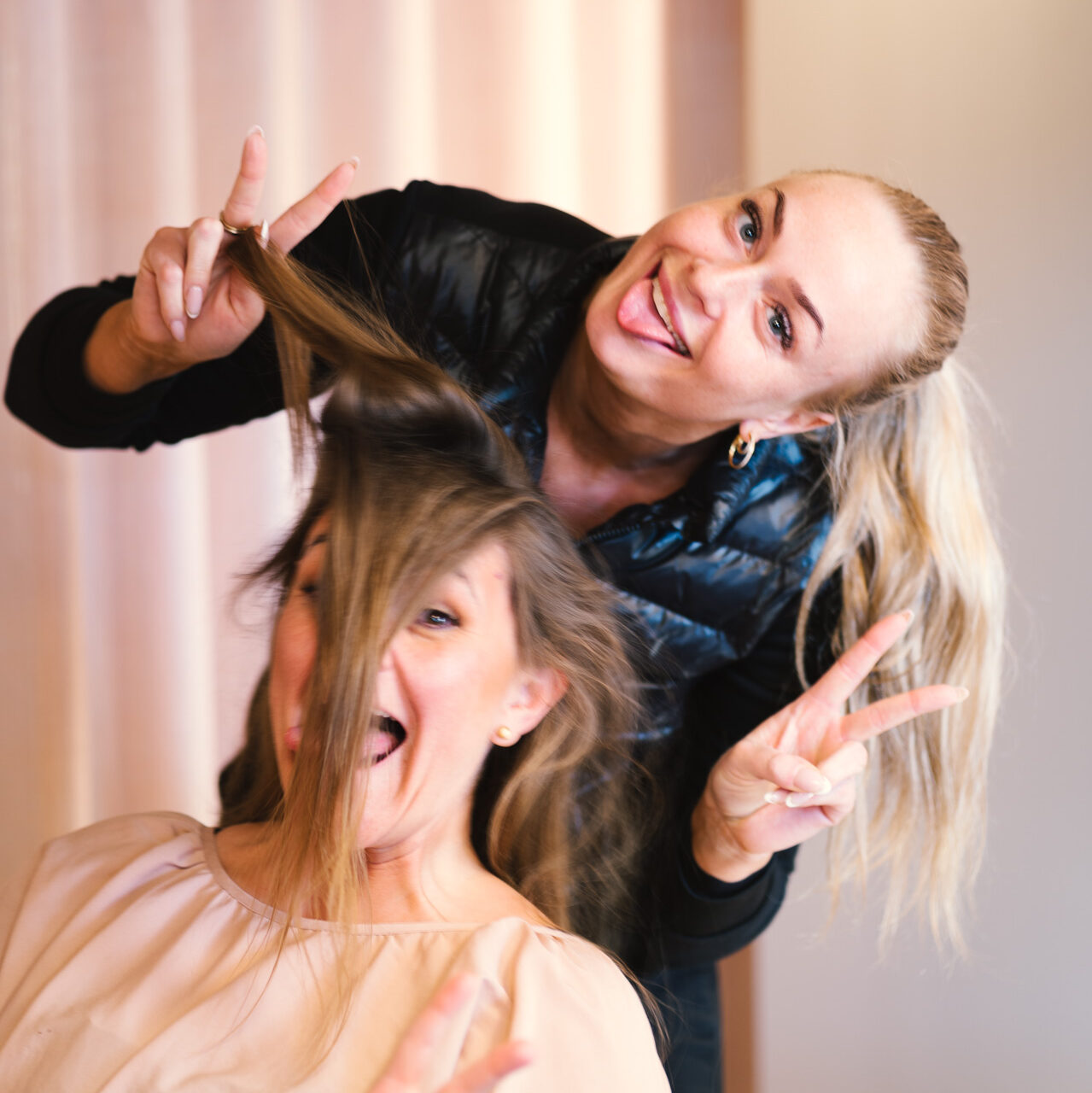 To kvinder makeupartisten og hårstylisten der laver sjove positurer, hvor den enehånd laver peace-tegn og den anden holder i håret