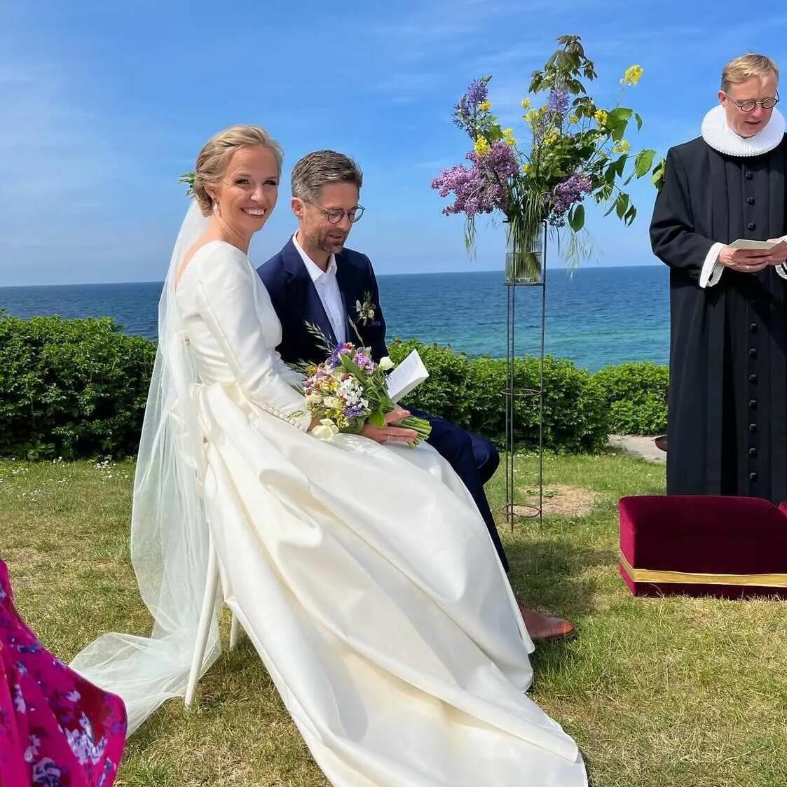 Brudepar ved udendørs vielse ved havet; bruden med naturlig brudemakeup, elegant opsat hår og langt slør.