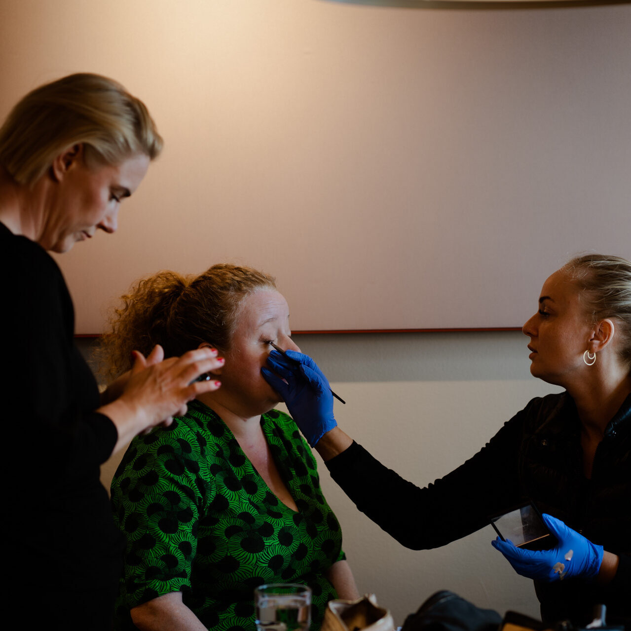 Makeupartist påfører makeup på kvinde i grøn kjole, mens en anden stylist forbereder produkter.