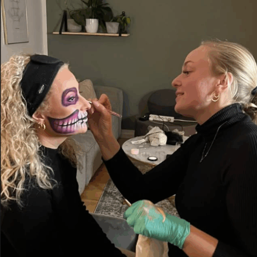 Makeupartist maler farverigt skull-facepaint i pink, lilla og hvid på kvinde med krøllet hår.