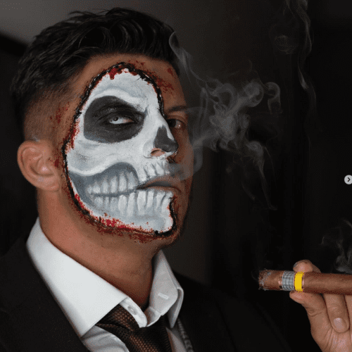 Mand med dramatisk skull facepaint, sorte øjenhuler, hvide knogledetaljer og røde sårkanter, mens han holder en cigar.